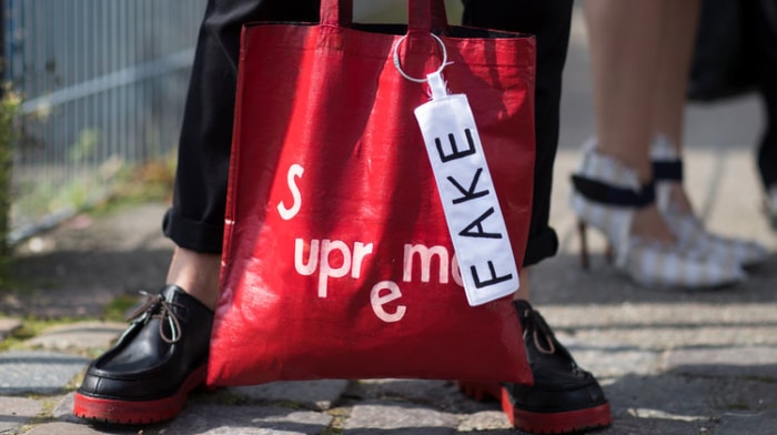 Bilde av en mann som holder en rød veske fra Supreme med en lapp der det står 'Fake'